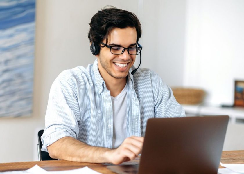 Happy successful confident young hispanic guy in headphones, call center employee or freelancer, working remotely on laptop, communicating via video communication with employees or client, smiling