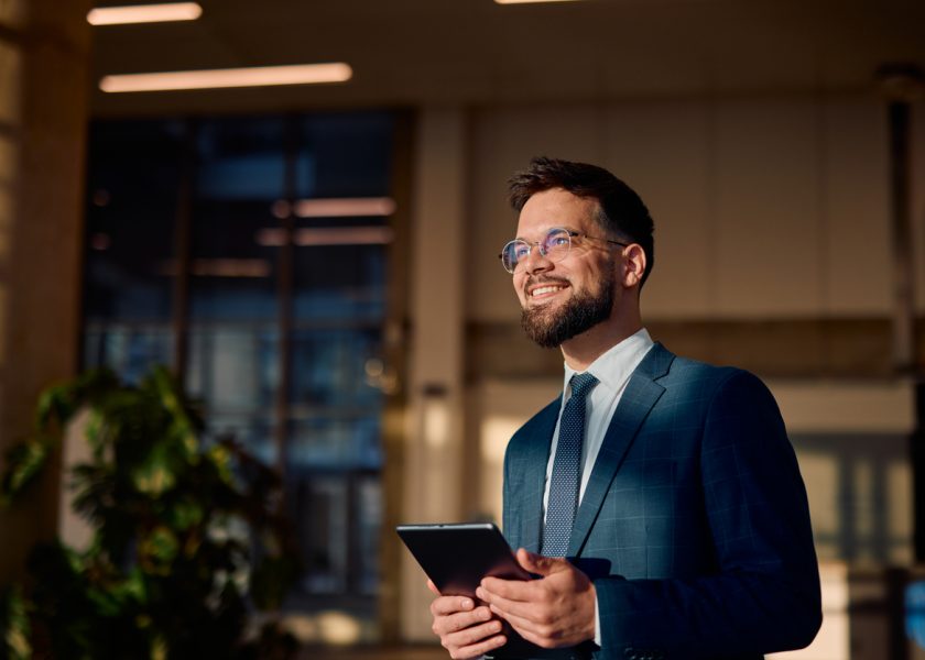 Smiling businessman holding tablet and looking up in office lobby